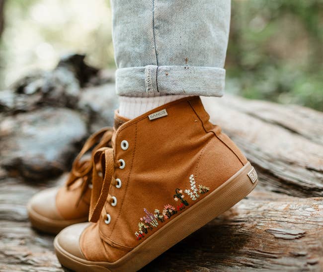 a model wearing a tan canvas hightop sneaker with floral embroidery along the side 
