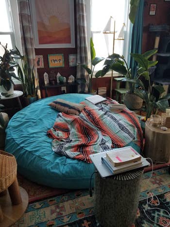 Cozy, bohemian-style room with a large bean bag covered by a patterned blanket, surrounded by plants and books on tables