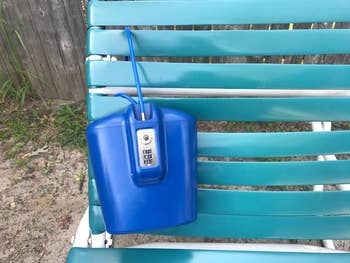 Portable bluish lockbox with a operation dial and cablegram sits connected a striped platform seat adjacent a woody fence