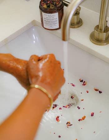 Person washing hands over sink with flower petals, near a jar of product, possibly linked to a beauty article