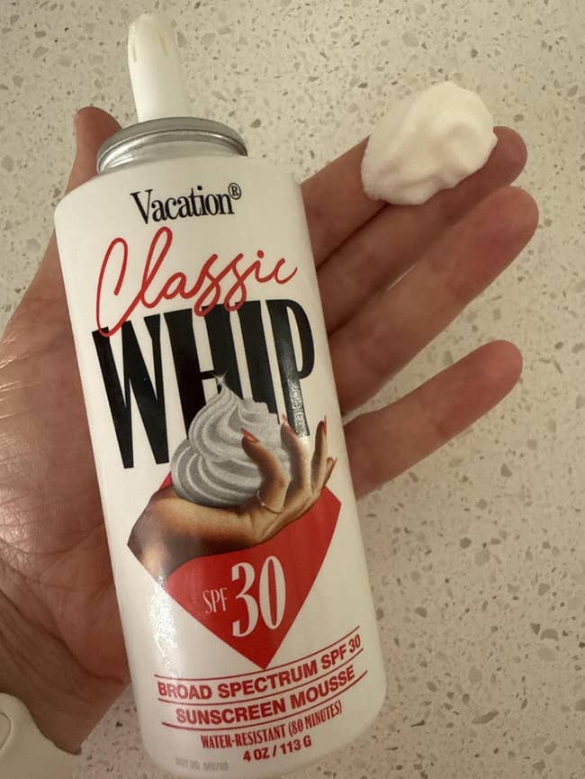Hand holding a can of Vacation Classic Whip SPF 30 sunscreen mousse with a dollop on the finger