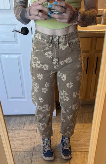 Person wearing floral-patterned pants, a cropped top, and bluish sneakers takes a reflector selfie, showing a casual and trendy outfit