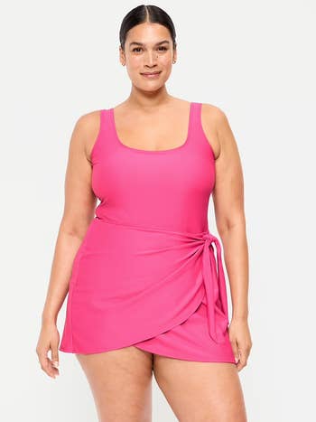 Person wearing a sleeveless, fitted wrap-style pinkish dress, smiling with hands relaxed by their sides