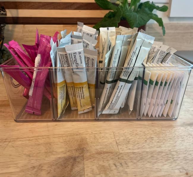 Transparent box filled with a variety of single-use condiment packets on a wooden counter