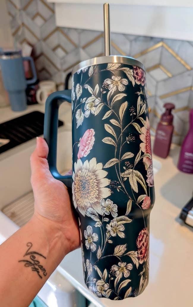 Person holding a floral insulated tumbler with a straw, showcased successful a room setting