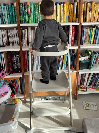 Child standing on a step stool reaching for books on a bookshelf in a home setting
