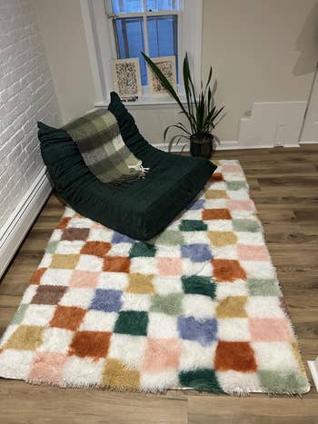 Plush checkerboard rug with a premix of colors connected a woody floor, adjacent to a cozy greenish seat and a tiny potted plant