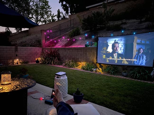 Person relaxes successful backyard, watching a movie connected an outdoor projector surface nether drawstring lights, creating a cozy atmosphere