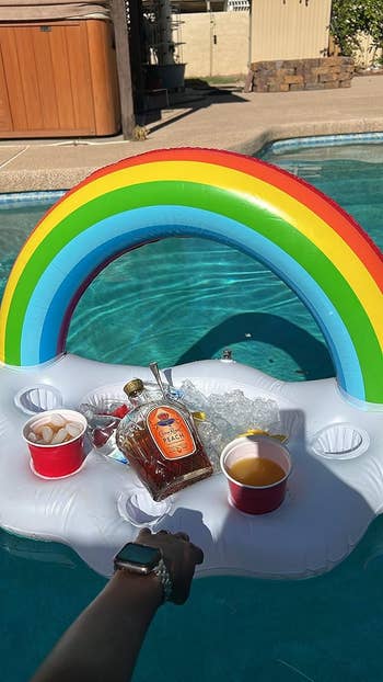 A pool float shaped like a rainbow holds a bottle of Jack Daniel's Apple Whiskey, ice, and red cups filled with drinks. A smartwatch-wearing hand is visible