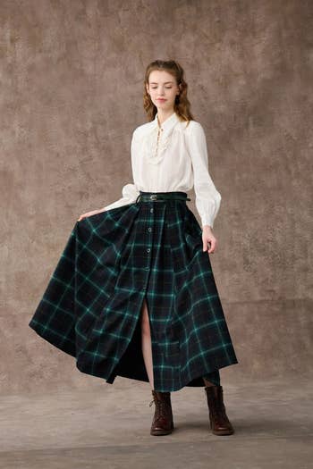 model wearing the green and blue plaid skirt with a white top and holding one side of the skirt out to show the slit at the bottom