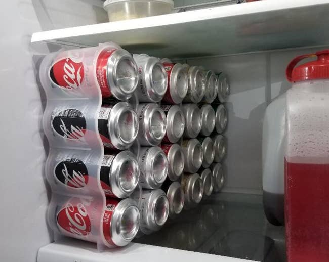 A set of four vertically stacked soda cans in a fridge held by a clear plastic container 