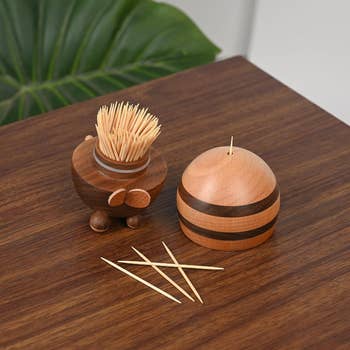 Wooden toothpick holders, one open revealing toothpicks and one closed, with scattered toothpicks on a wooden table