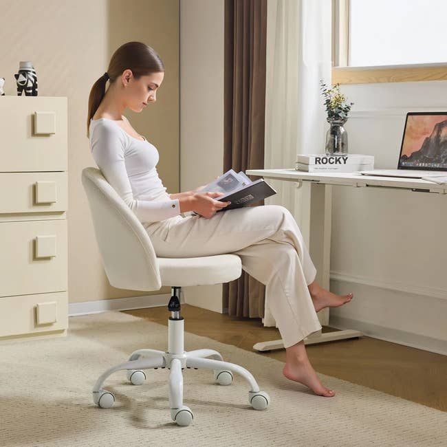 A model in a white top and light pants sits on a modern swivel chair in a home office setting, reading a magazine