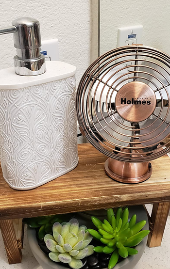 a reviewer photo of a small copper fan sitting on a wooden stand next to a hand soap bottle