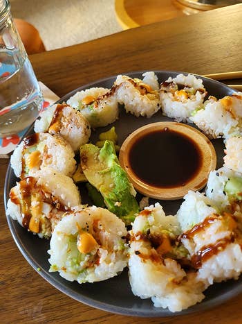 Plate of sushi rolls drizzled with sauce, with avocado and a vessel of soy sauce