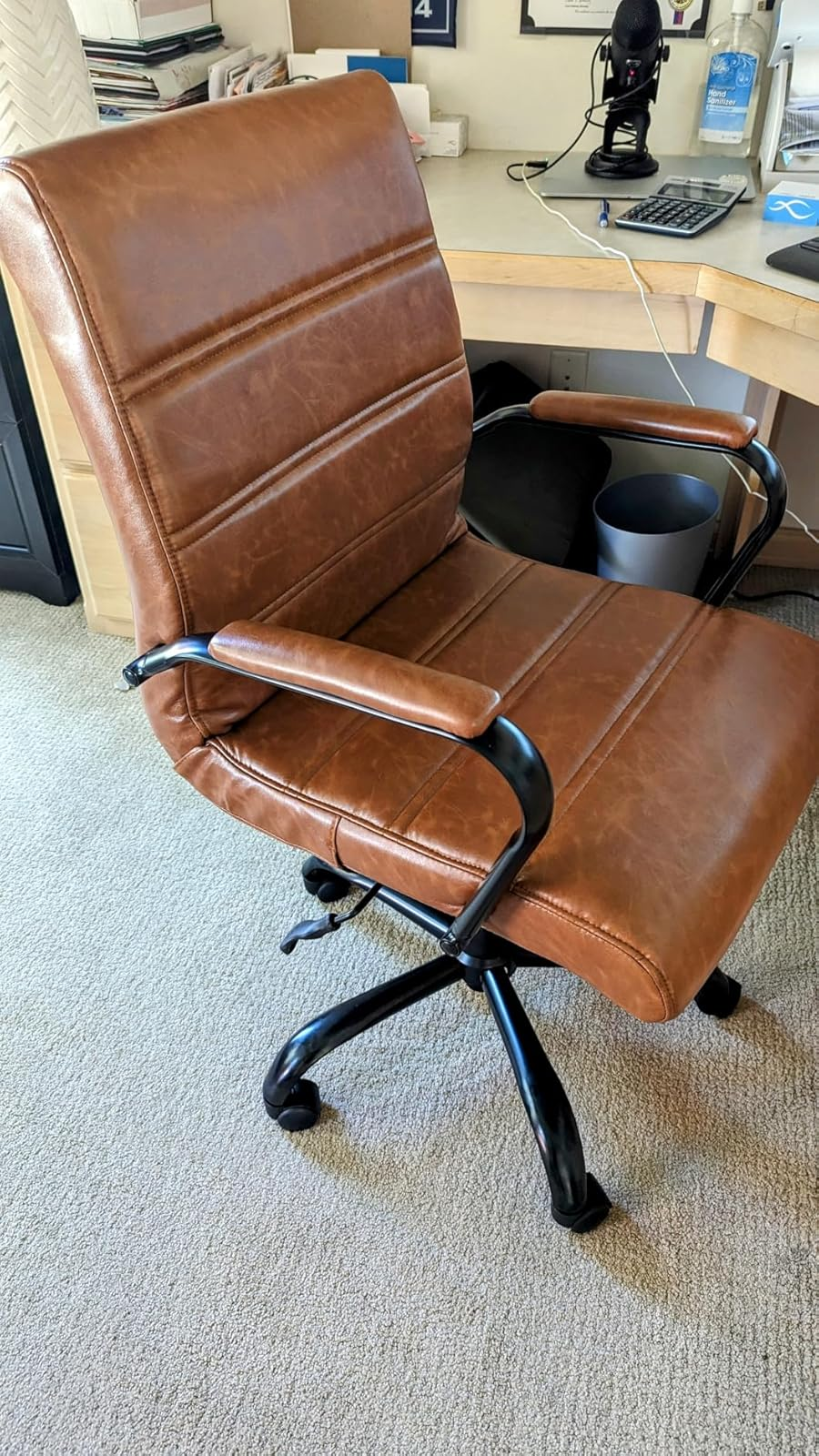 reviewer photo of a brown leather office chair with armrests and black metal base on a beige carpet in a home office setting