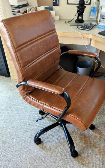 reviewer photo of a brown leather office chair with armrests and black metal base on a beige carpet in a home office setting