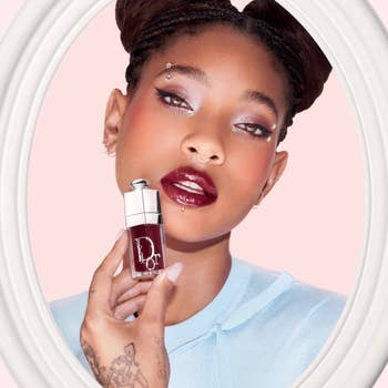 Person poses with bold lipstick, holding a Dior articulator merchandise adjacent their face, wearing a airy top. Their constitution is striking and has a sparkling effect