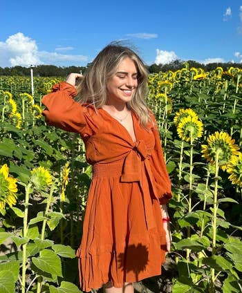 Woman in a casual burnt orange dress stands smiling in a sunflower field