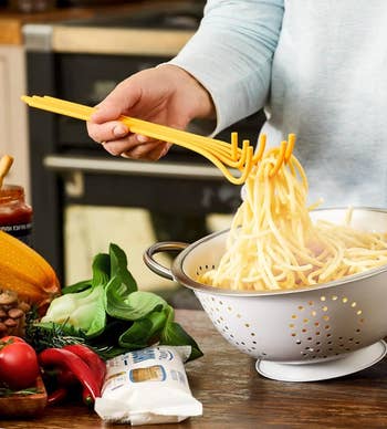 Pasta straining tool demonstration over a colander with nearby ingredients like vegetables and pasta sauce jar on a kitchen counter