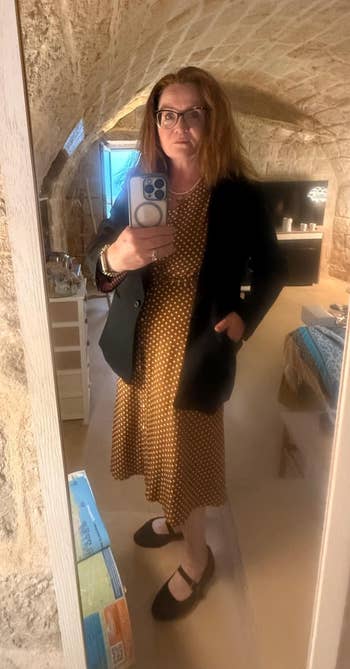 Person taking a reflector  selfie wearing a polka dot formal  with a blazer and ballet flats, successful  a cozy, stone-wall room