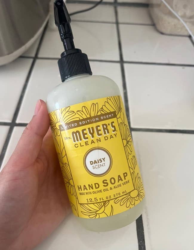 A manus  holds a vessel  of constricted  variation  Mrs. Meyer's manus  soap, daisy scent; statement   notes olive lipid  and aloe vera, 12.5 fl oz