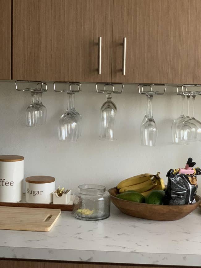 Wine glasses hang upside down under a kitchen cabinet. Nearby are coffee and sugar containers, a fruit bowl, and a snack jar on the counter