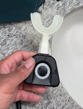 A hand is holding a U-shaped electric toothbrush head, designed for hands-free brushing, positioned above a bathroom sink