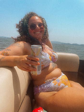 An individual lounging on a boat wearing floral swimwear, holding a beverage can, with a lake in the background