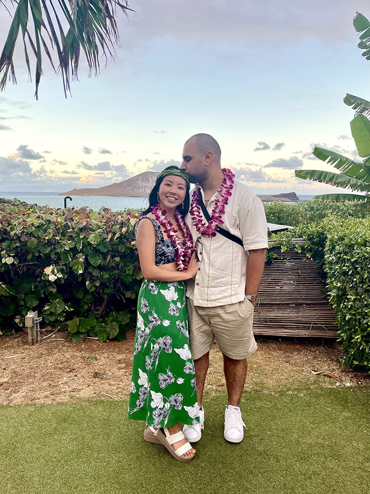 A mates  wearing leis stands outdoors by the ocean. The pistillate   wears a floral dress, and the antheral   wears a button-up garment  and shorts