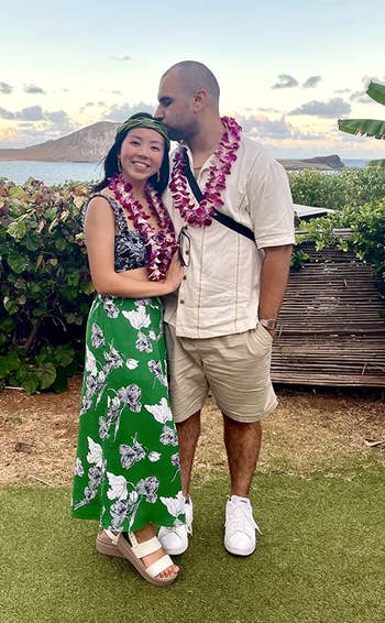 A mates wearing leis stands outdoors by the ocean. The pistillate wears a floral dress, and the antheral wears a button-up garment and shorts