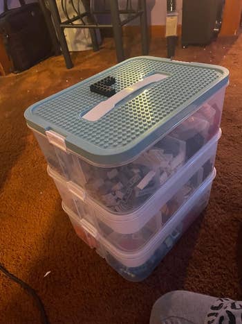 Kotak penyimpanan plastik bening bertumpuk berisi berbagai macam potongan LEGO, dilengkapi pegangan kokoh pada tutupnya, diletakkan di atas lantai berkarpet