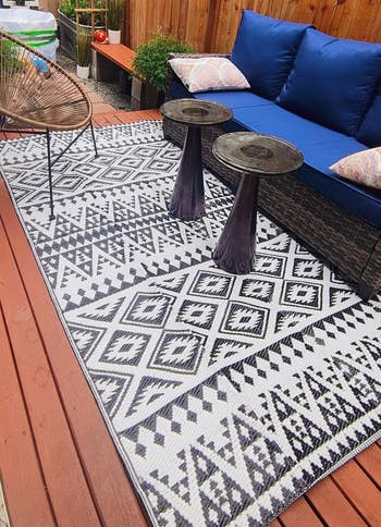 Outdoor patio with geometric-patterned rug, wicker sofa with plush cushions, woody fencing, and tiny tables. Cozy setup for relaxing oregon entertaining