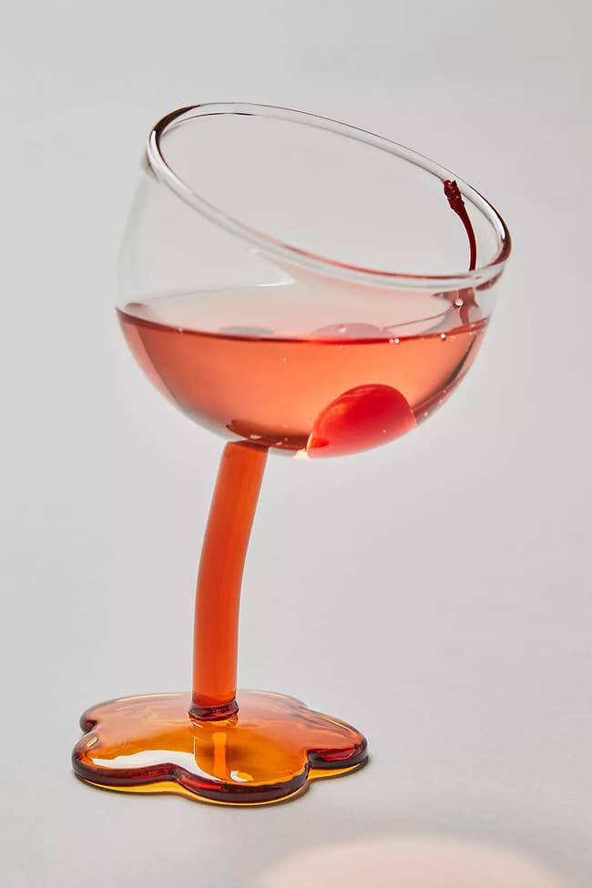 A clear wine glass with an orangey-red tilted stem and a base shaped like a flower 