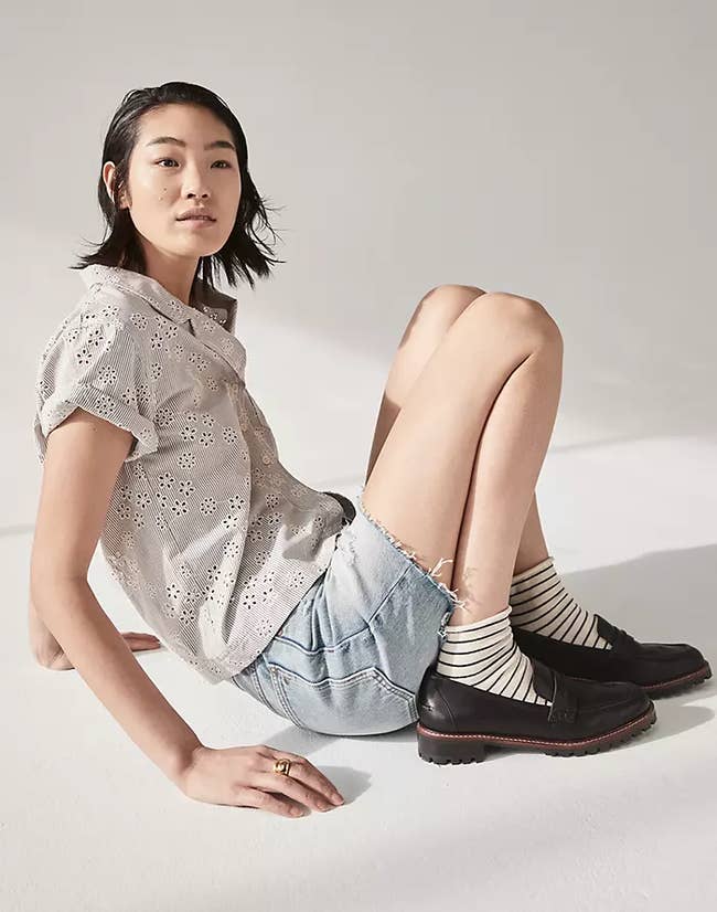 Model sitting on the ground with legs curled up to showcase the loafers in black. They also have a thin brown line around the top of the sole where the stitching is