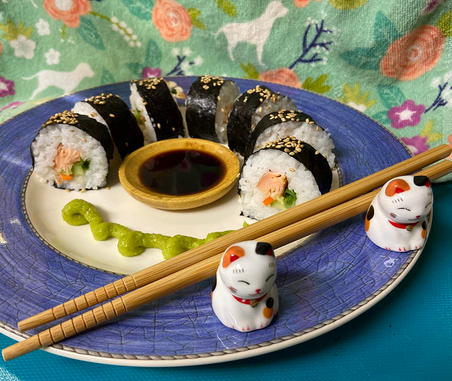 Sushi rolls with salmon and avocado, served on a plate with soy sauce and wasabi, chopsticks, and cat figurines; floral background