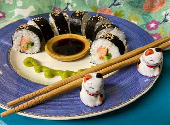 Sushi rolls with salmon and avocado, served on a plate with soy sauce and wasabi, chopsticks, and cat figurines; floral background