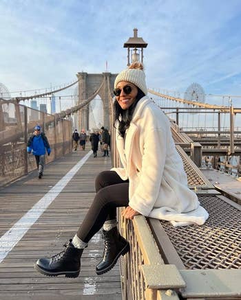 reviewer successful  a cozy coat, hat, and boots sits connected  Brooklyn Bridge railing, smiling for a shopping-themed article