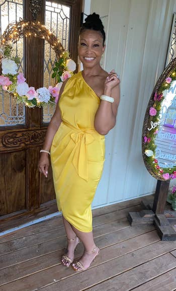 Woman in chic yellow mid-length dress with tie waist detail and jeweled sandals, posing on a porch for a stylish look