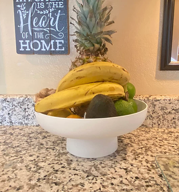 A small white elevated fruit bowl with bananas and apples in it 