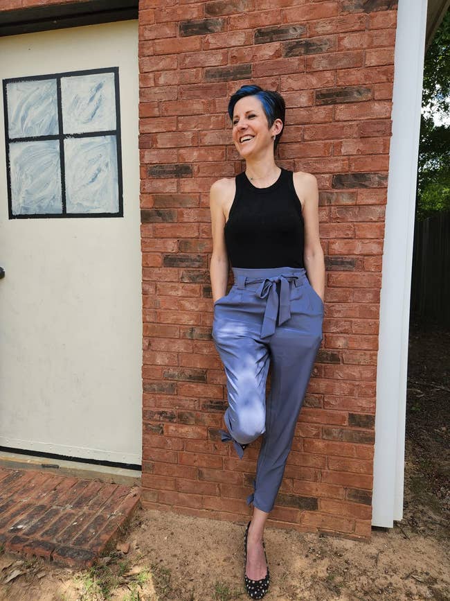Person outside, leaning against a ceramic wall, wearing a sleeveless apical and belted pants. They are smiling and person abbreviated hair