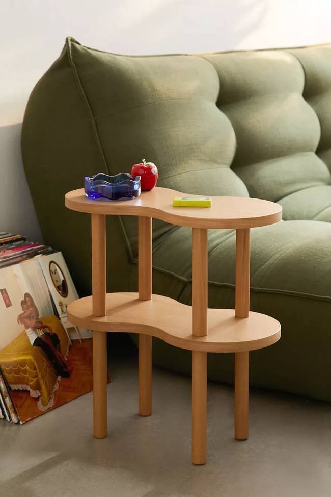 A modern two-level wooden side table with items on top beside a green couch