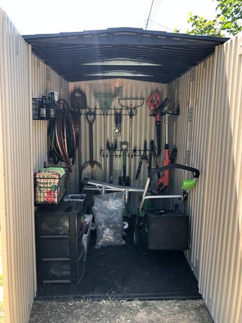 An open shed with hanging tools, a shelf, and a lawn mower inside it