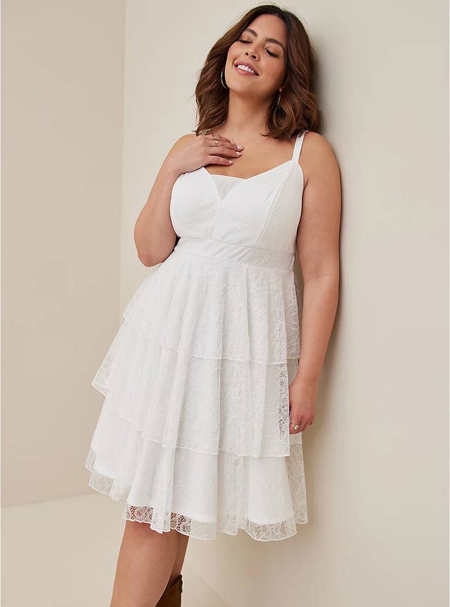 Model wearing white sleeveless knee-high tiered lace dress in white with a sweetheart neckline standing in front of a white wall wearing shin tall brown shoes