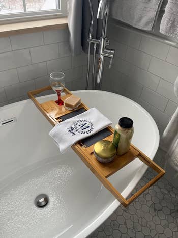 Wooden bath tray on a tub with a candle, body wash, wine glass, and scrub brush, promoting a relaxing shopping experience
