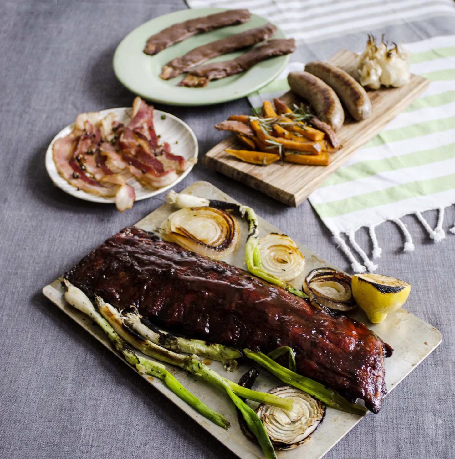 Plates and serving platters across a gray table with ribs, grilled vegetables, bacon, and sausage