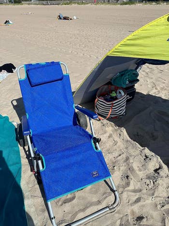 Beach chair under a sunshade with a striped bag, on sandy beach with people relaxing in background