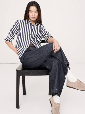 Model seated, wearing a striped button-up garment and wide-leg jeans with loafers. Fashionable, casual style