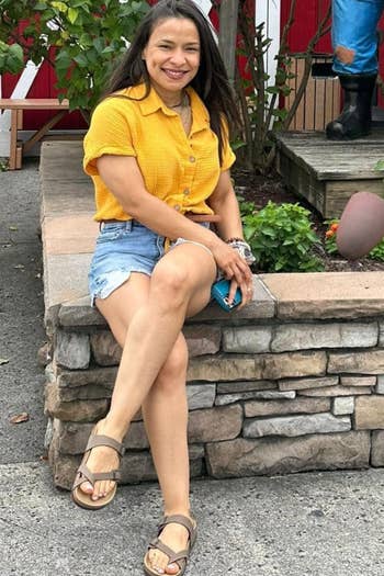 the reviewer sitting on a stone ledge wearing a yellow shirt, denim shorts, and sandals, smiling at the camera