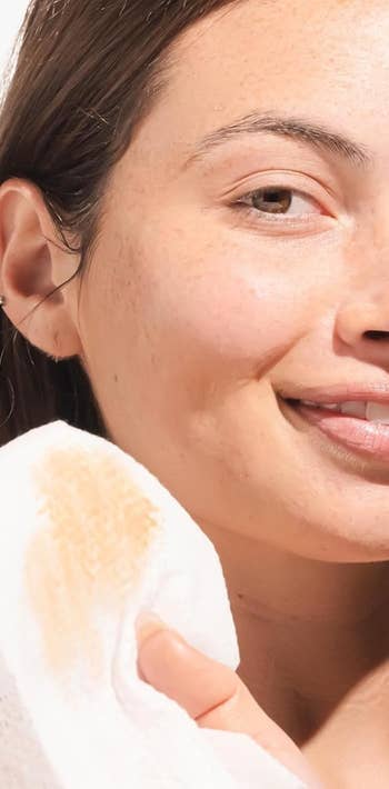 Person smiling portion holding a constitution remover pad adjacent to her face, showcasing a skincare routine